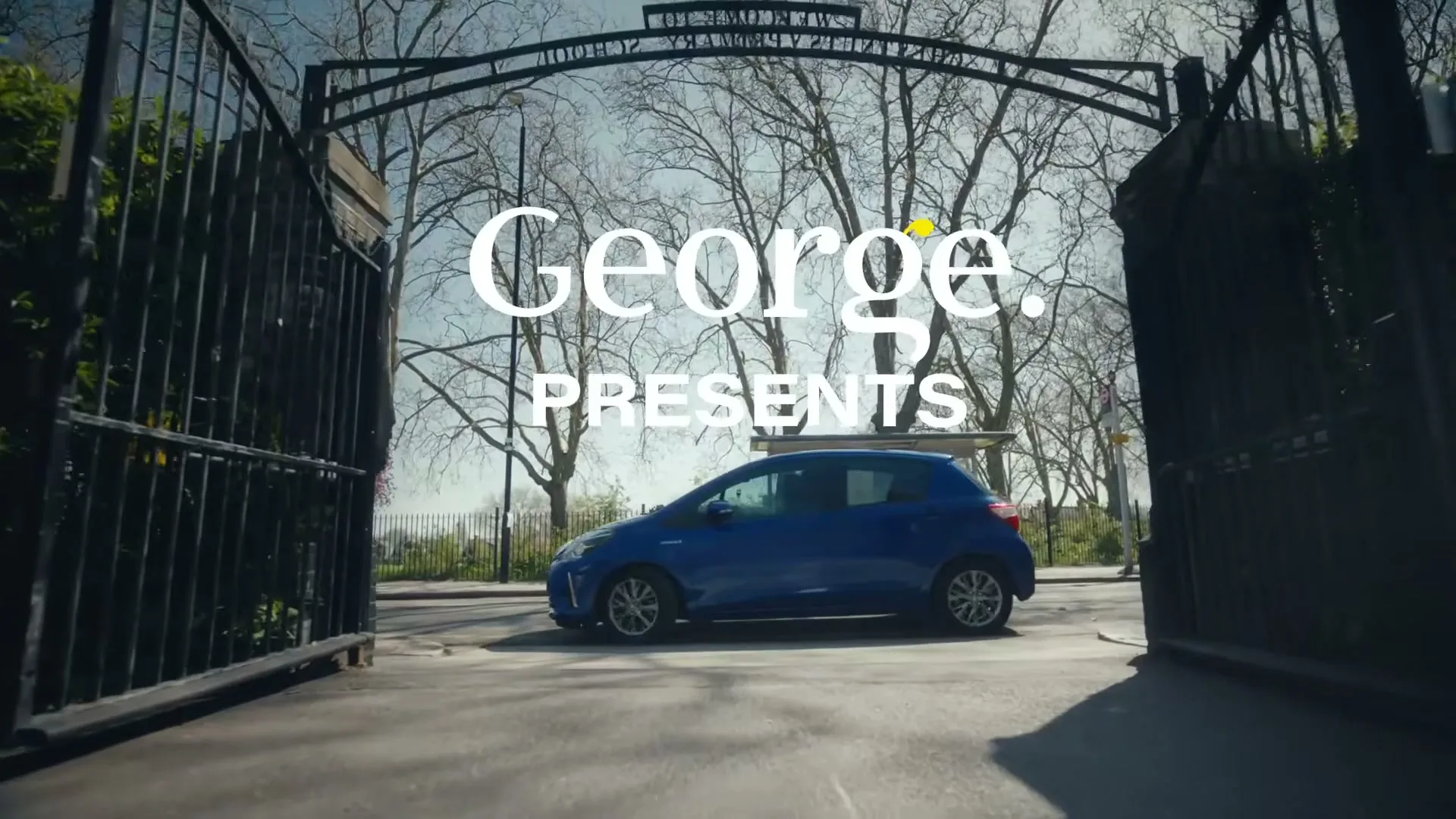 A blue car drives through an open ornate black gate on a sunny day, with the words "George. PRESENTS" overlaid on the screen.