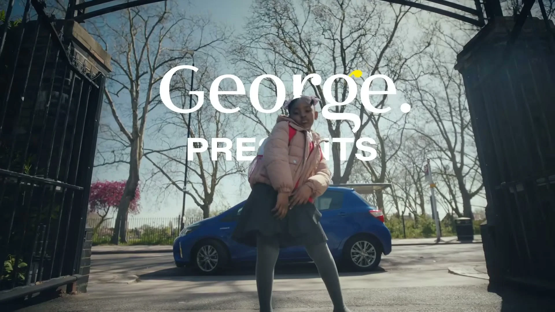 A young girl in a pink puffer jacket and school uniform dances confidently in front of a blue car, framed by large iron gates on a sunny day.