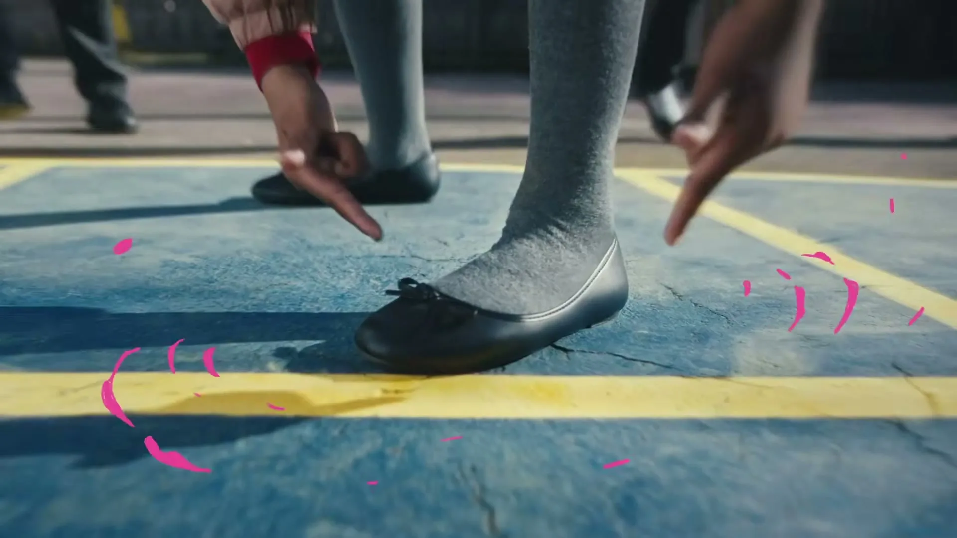 A child's feet in black ballet flats and grey socks are centered on a blue and yellow painted ground, with other children's feet in the background.
