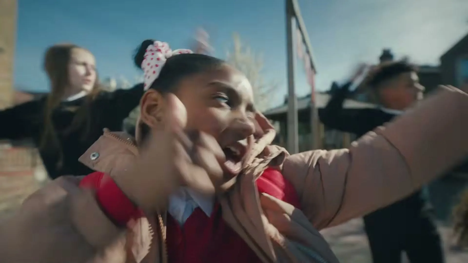 A young Black girl in a school uniform and a puffer jacket dances energetically with other children in a sunny outdoor setting.