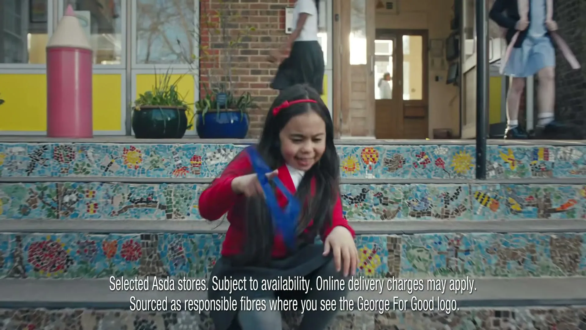 A young girl with long dark hair and a red cardigan sits on colorful mosaic steps outside a school, playing with a blue toy.