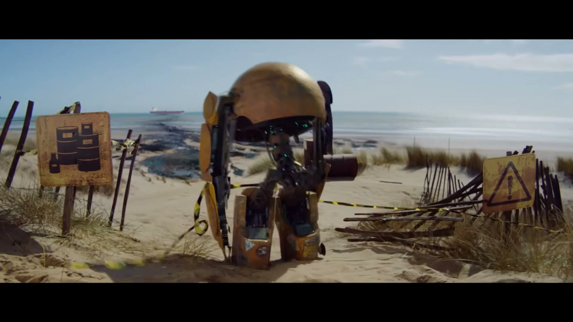 A small, rusty, yellow robot with a domed head walks on a sandy beach with warning signs and the ocean in the background.