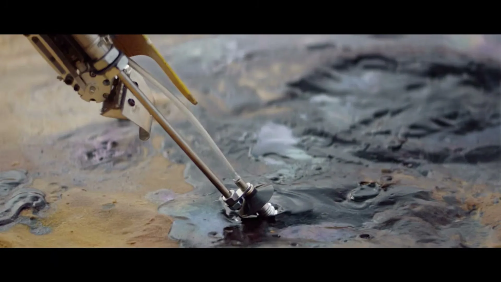 A robotic arm with a small nozzle drips a dark, viscous liquid onto a surface with similar liquid puddles, creating ripples.