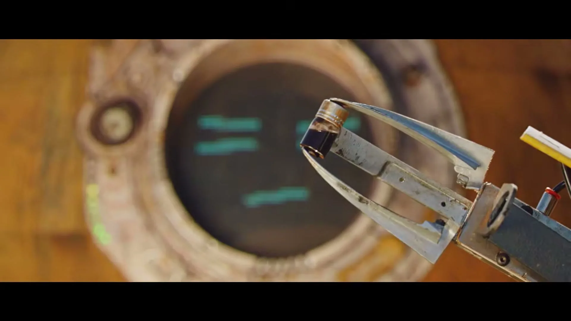 A robot's claw holds a small vial in front of its screen face, which displays a pixelated, sad expression.