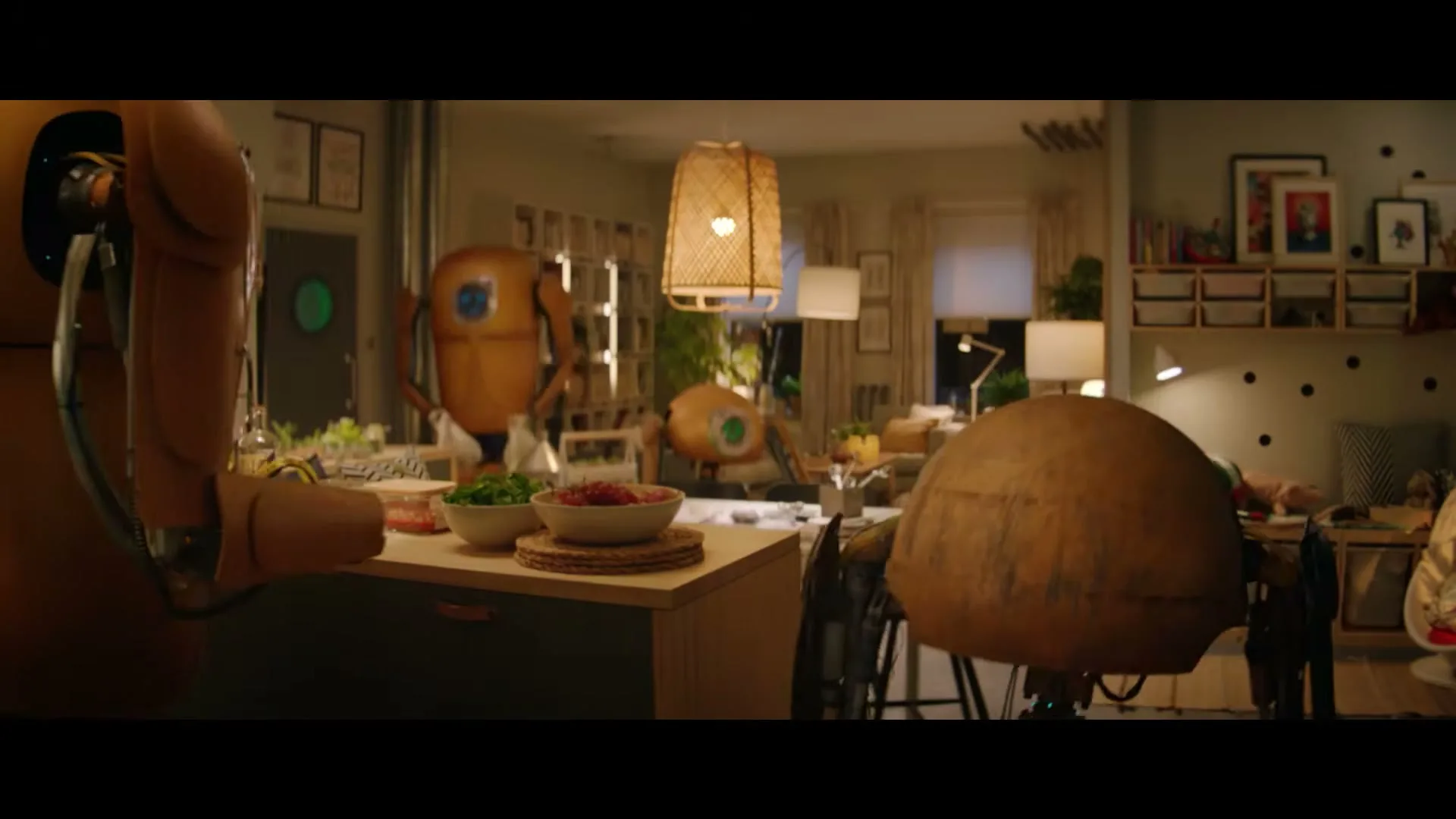 A group of small, brown, spherical robots with large eyes are seen in a cozy, well-lit living room, with one robot in the foreground on a kitchen island.