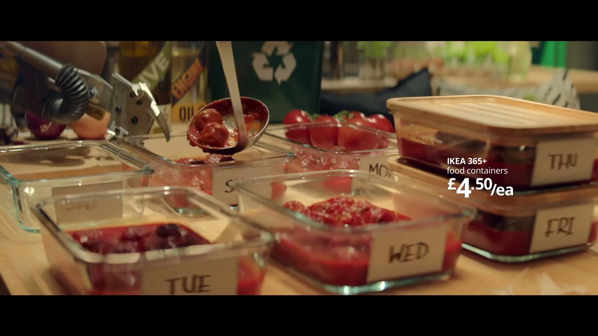 A robot arm uses a ladle to scoop meatballs and sauce from a can into glass food containers labeled with days of the week on a wooden counter.