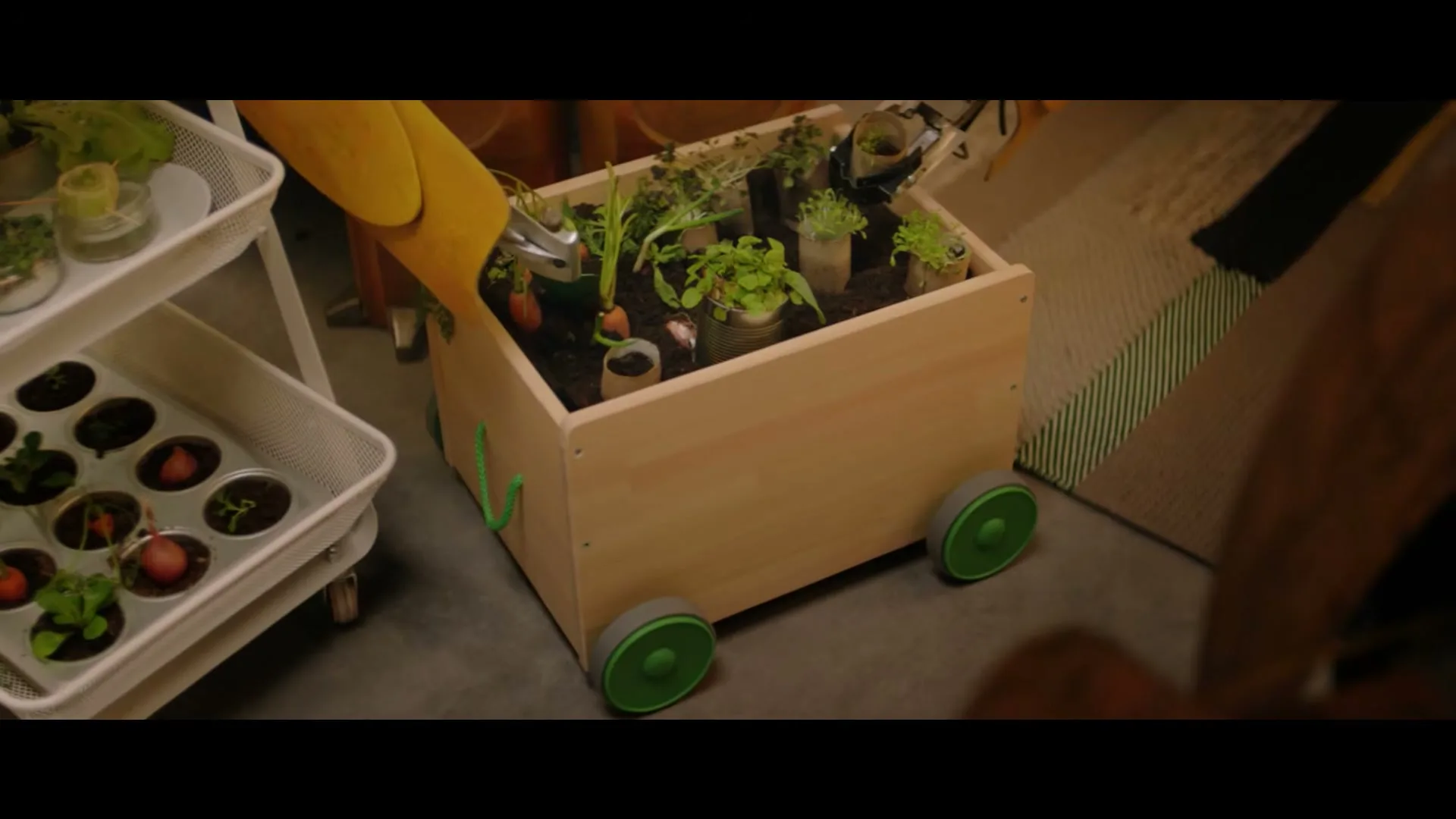 A yellow robot arm tends to small plants growing in a wooden cart with green wheels, next to a white rolling shelf with more plants.