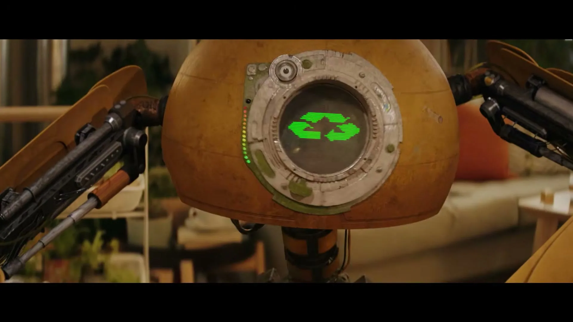 A small, round, yellow robot with mechanical arms displays a green recycling symbol on its screen in a living room.