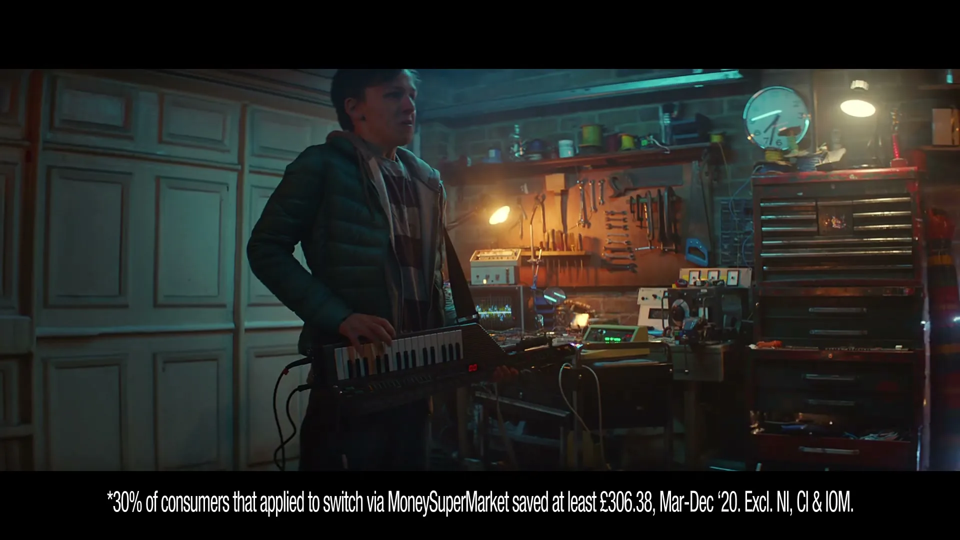 A man in a green jacket plays a keytar in a cluttered garage, looking around with a surprised expression.