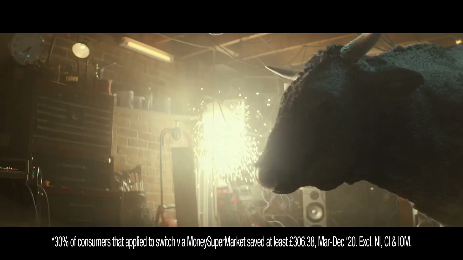 A large, dark bull stands calmly in a cluttered garage as sparks fly from a welding machine in the background, unfazed by the chaos.