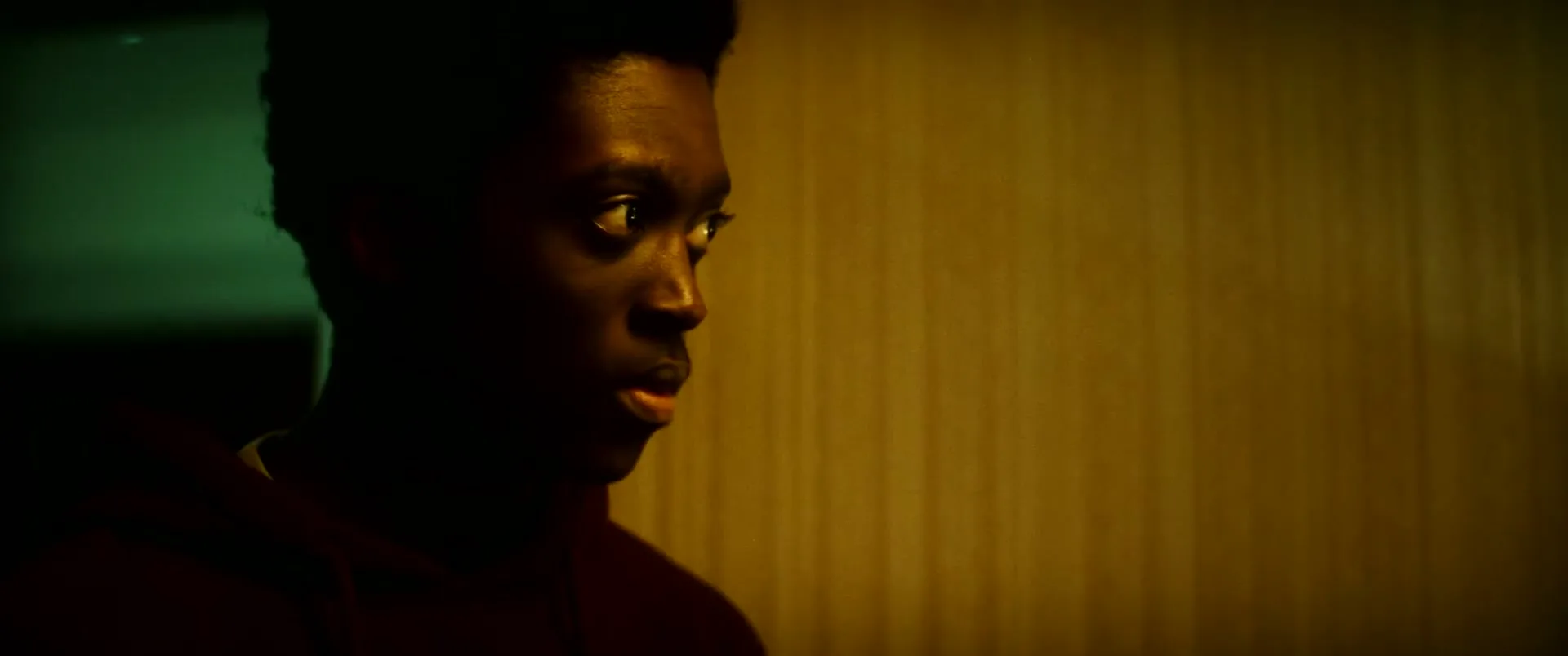 A young Black man in a dark hoodie looks around nervously in a dimly lit hallway, his face partially obscured by shadow.