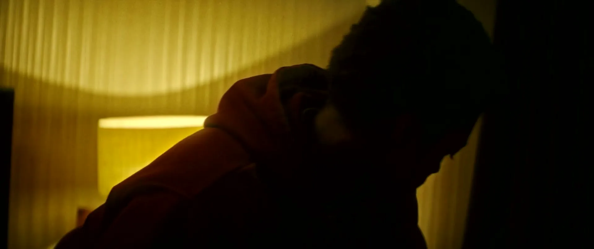 A young Black man in a red hoodie is seen in a dark room, leaning over something on the floor, with a lamp glowing in the background.