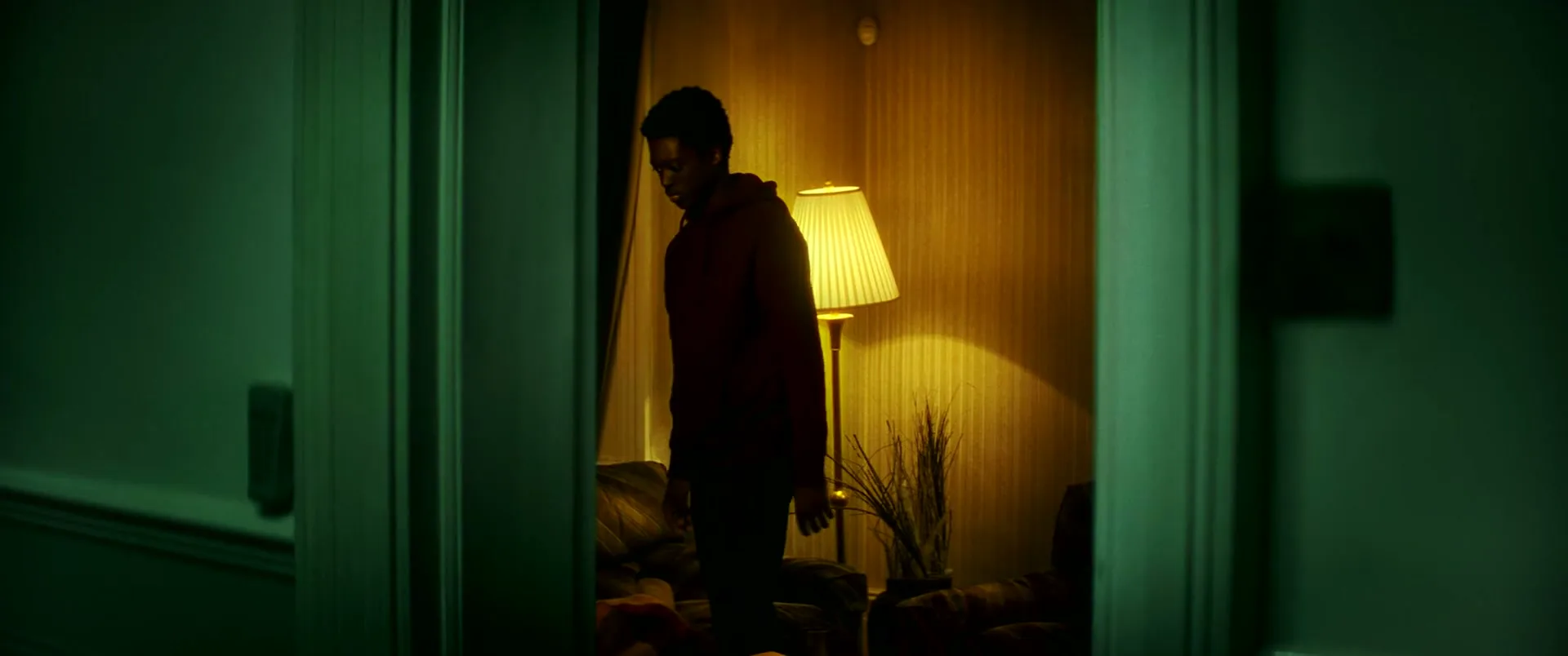 A young Black man in a dark hoodie walks into a dimly lit room, picks up a blanket, and then walks towards the camera.
