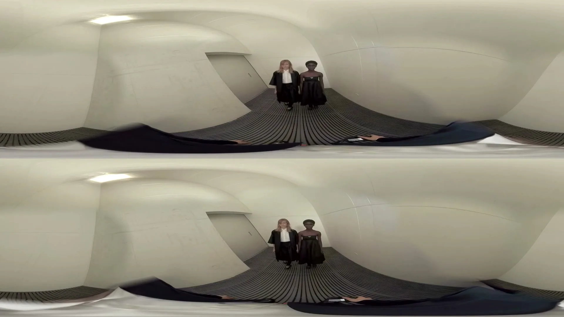 Two models, one blonde and one dark-haired, wearing black dresses, walk towards the camera and then stand back-to-back in a white, futuristic room.