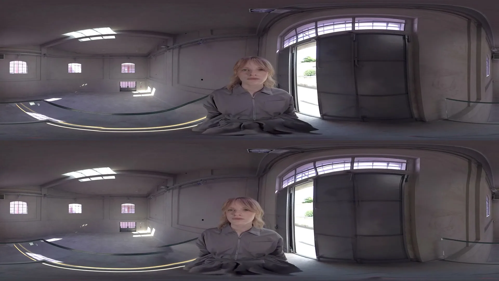 A young woman with blonde hair, wearing a gray jumpsuit, sits on the floor in a large, empty room with an open door to the outside.