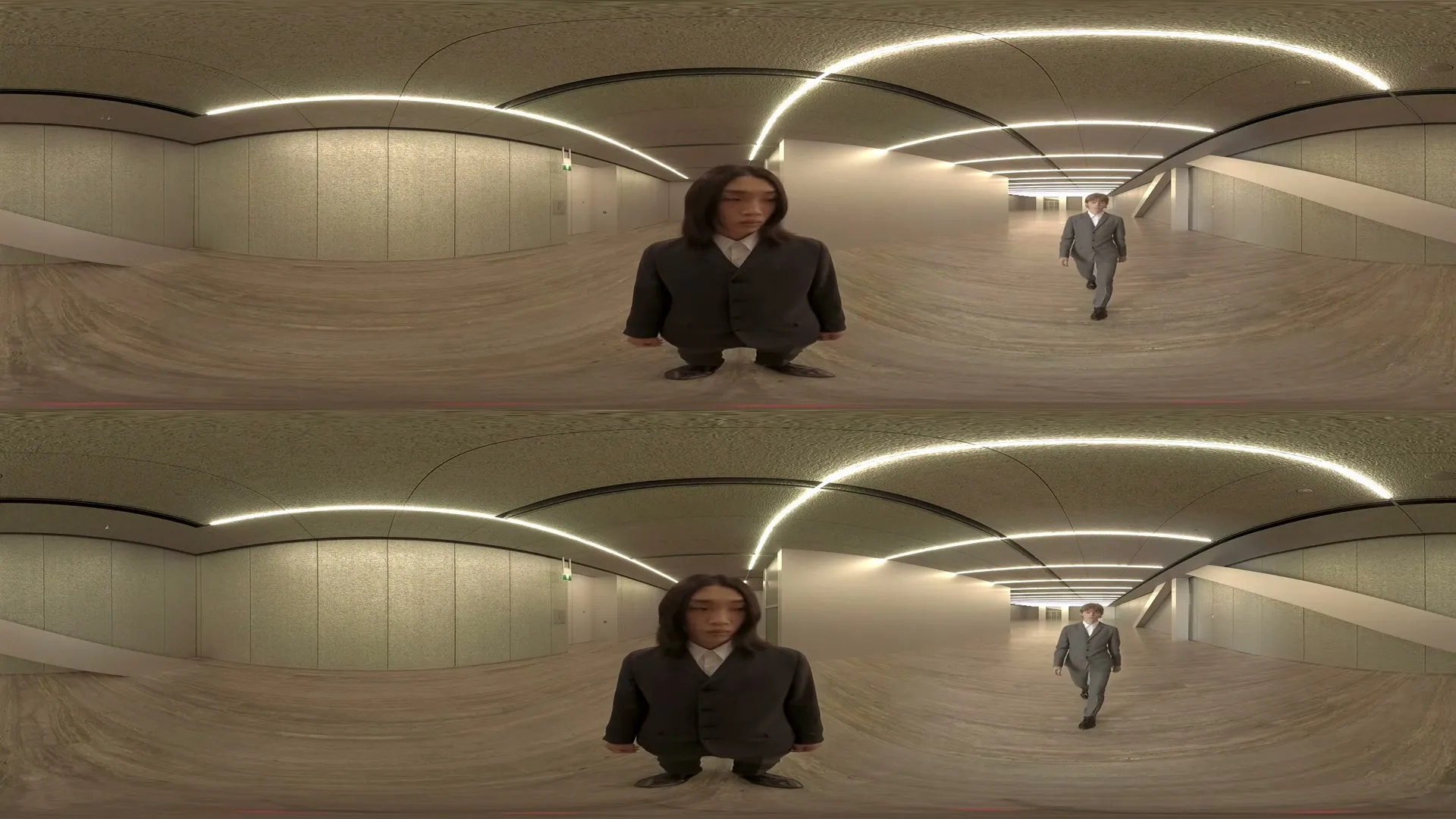 Two men in suits, one in the foreground and one in the background, walk towards the camera in a long, curved, well-lit hallway.