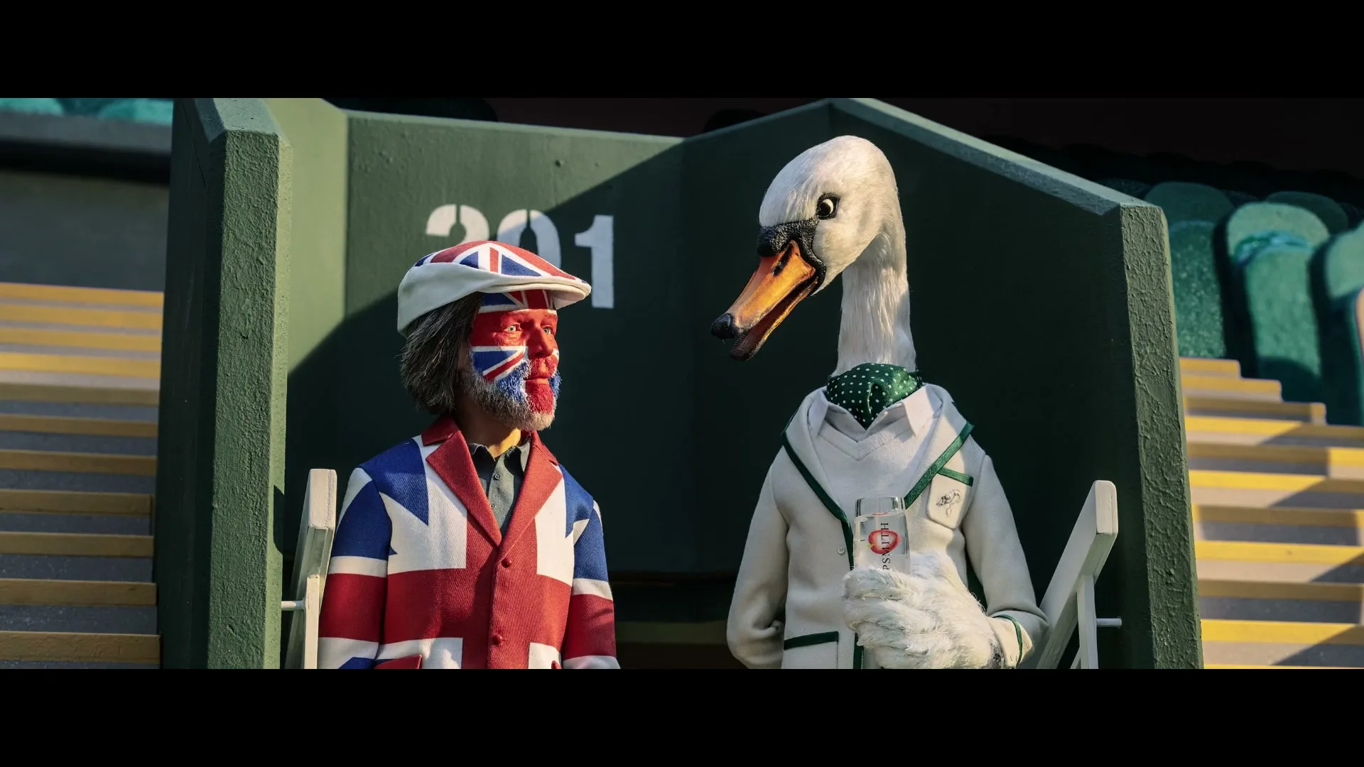 A man with a painted Union Jack face and jacket waves next to a swan in a tennis uniform, both sitting in stadium seats.