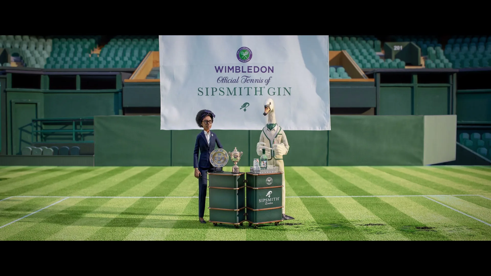A female puppet in a suit and a swan puppet in a tennis outfit stand next to a trophy and a gin bottle on a tennis court, with a banner behind them.
