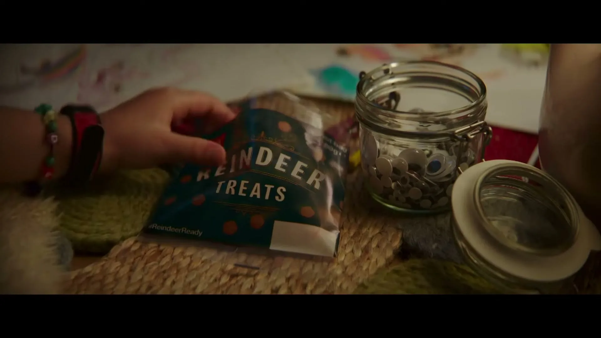 A child's hand reaches for a bag of "Reindeer Treats" next to a jar of googly eyes on a woven mat, suggesting a craft activity.