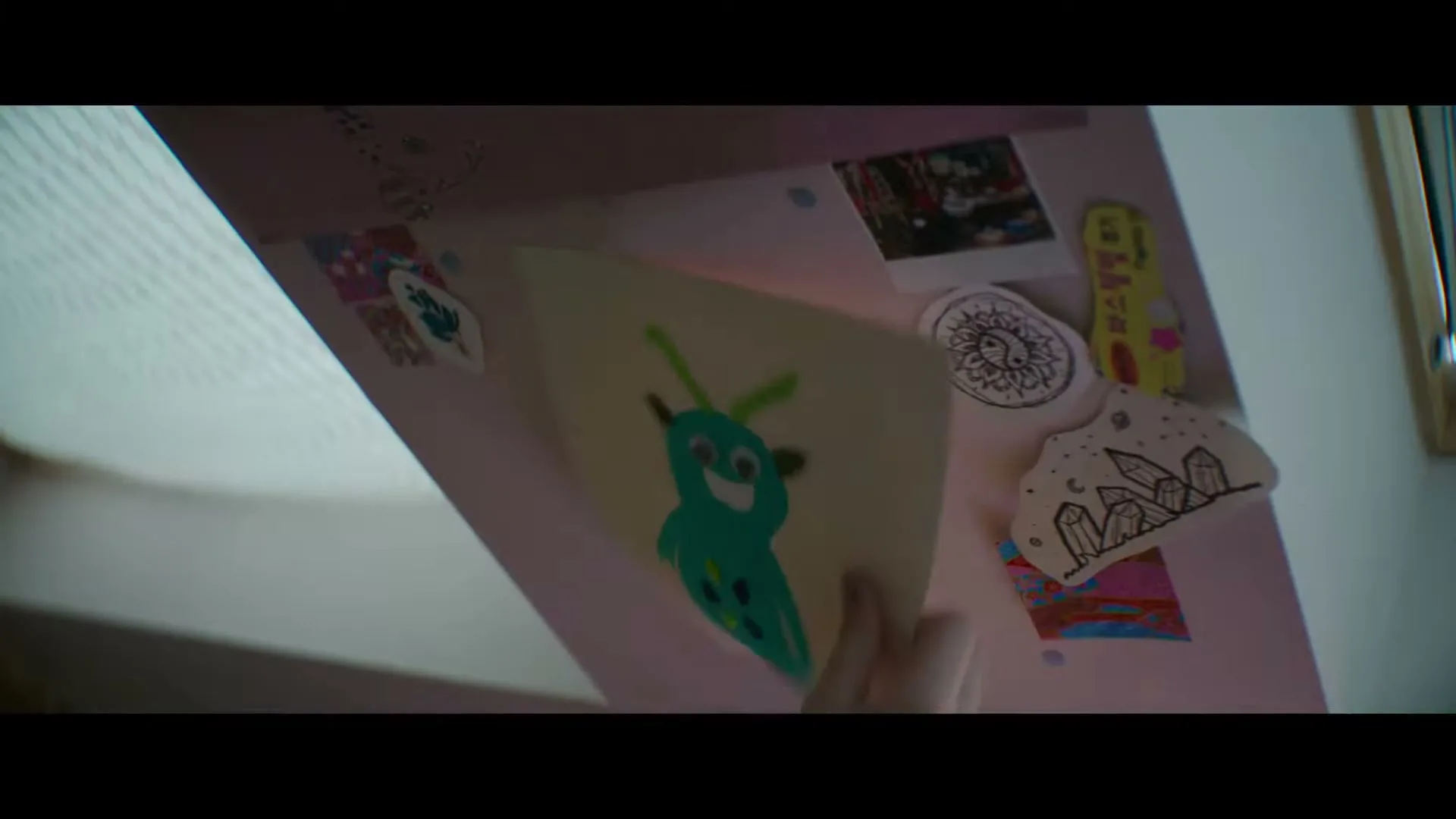 A child's hand removes a drawing of a blue monster from a pink board covered with other drawings and stickers.