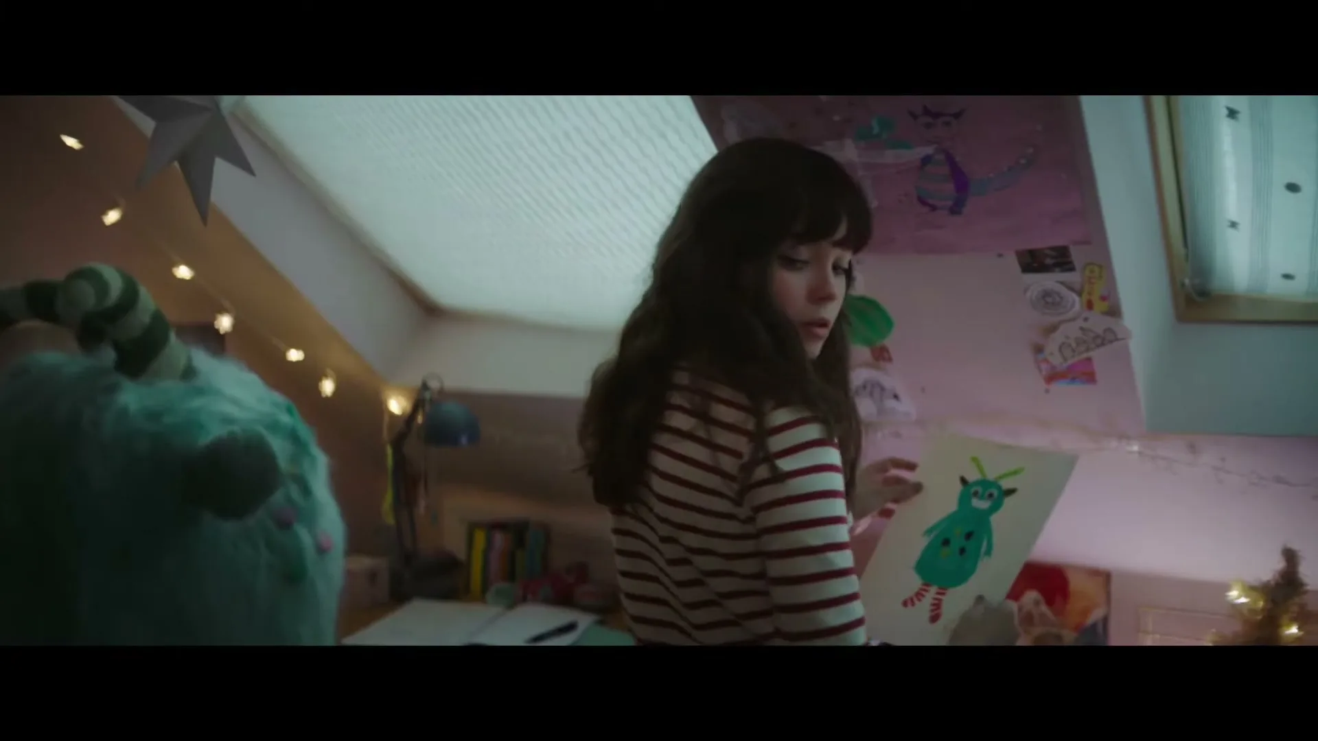 A young girl with dark hair in a striped shirt holds up a drawing of a green monster in her attic bedroom.