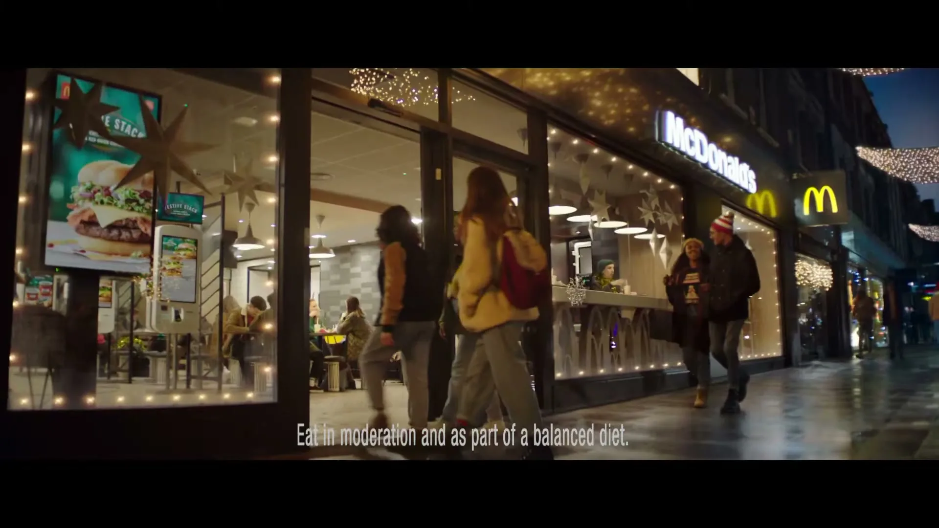 A wide shot of a McDonald's restaurant at night, with people walking past and inside, decorated with festive lights.