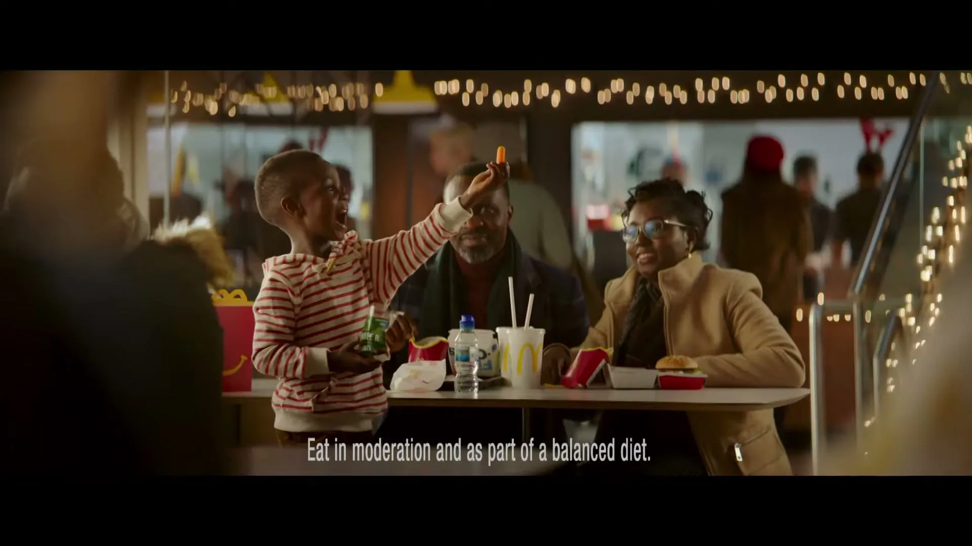 A young boy in a striped shirt holds a carrot stick up to a man's nose, while a woman smiles beside them at a McDonald's restaurant.
