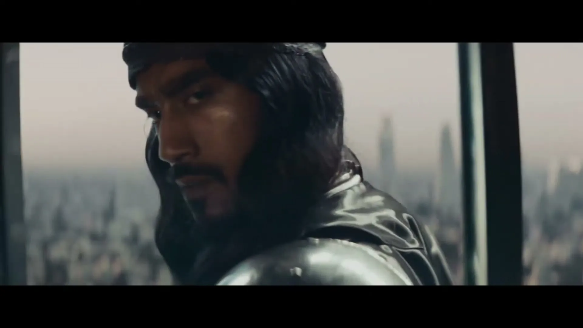 A man with long dark hair and a beard, wearing a dark headwrap and armor, looks over his shoulder with a serious expression, with a city skyline in the background.