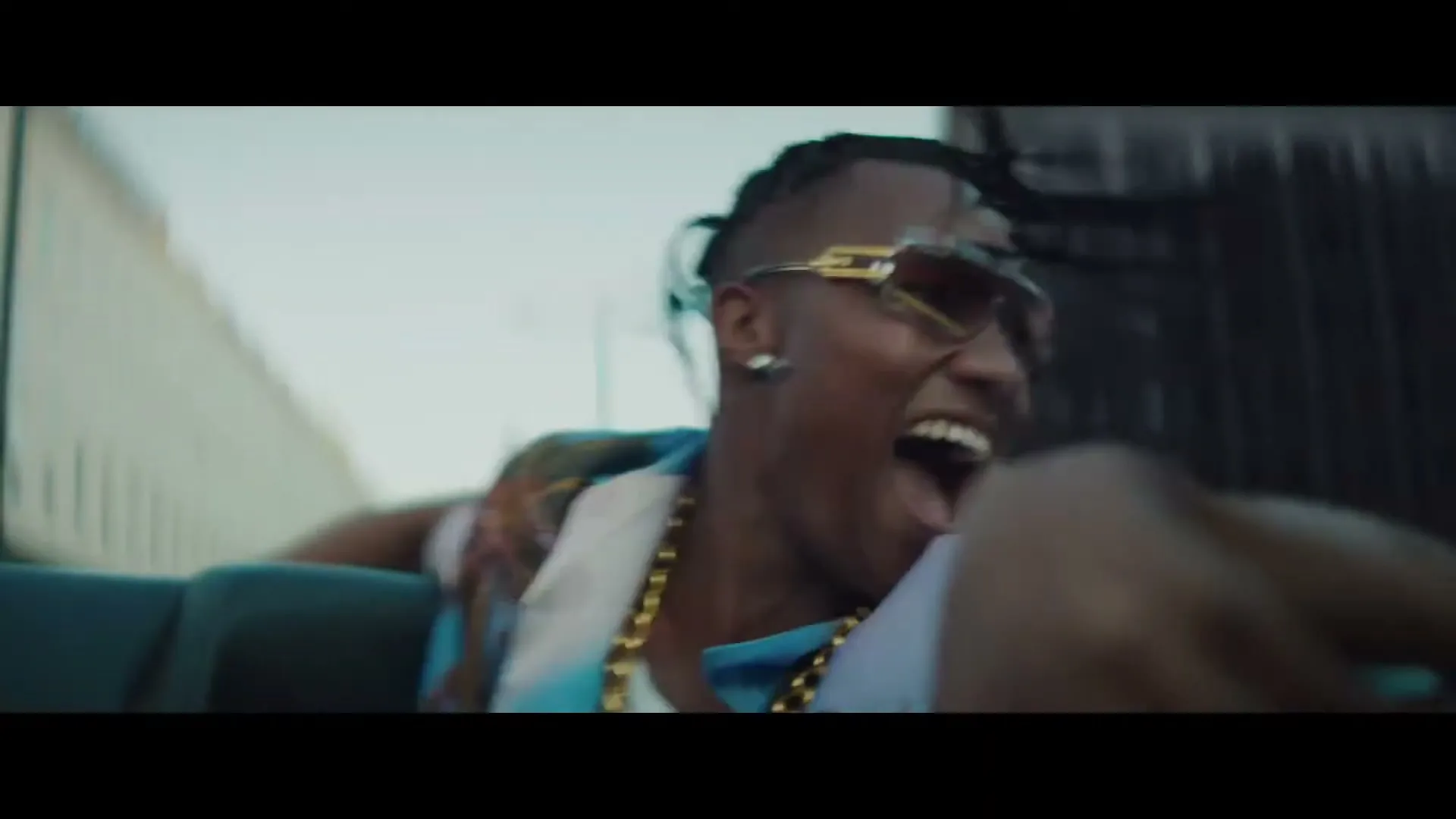 A young man with dreadlocks and sunglasses, wearing a gold chain and Hawaiian shirt, screams joyfully in the back of a moving car during the day.