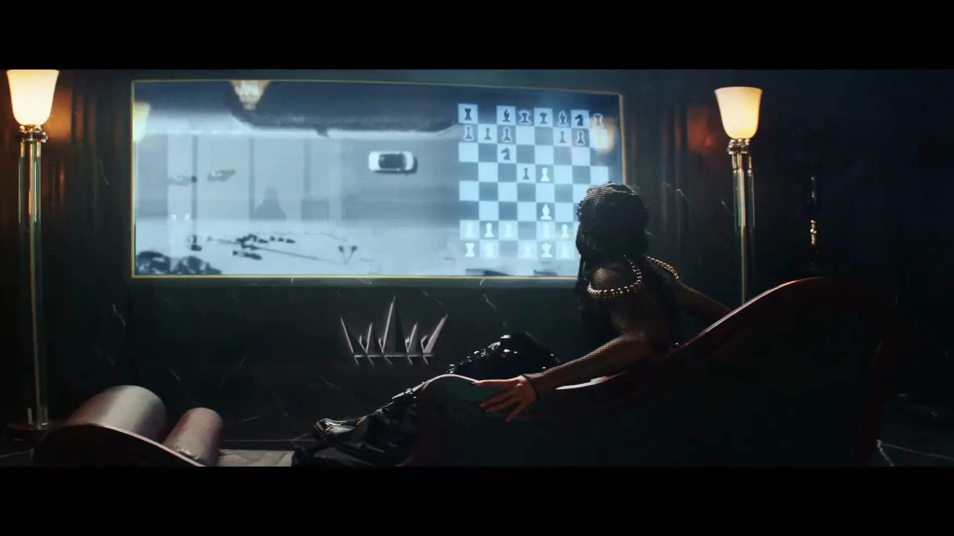 A person with braided hair and a dark, ornate outfit reclines on a chaise lounge, watching a large screen displaying a chess game and a desert landscape.