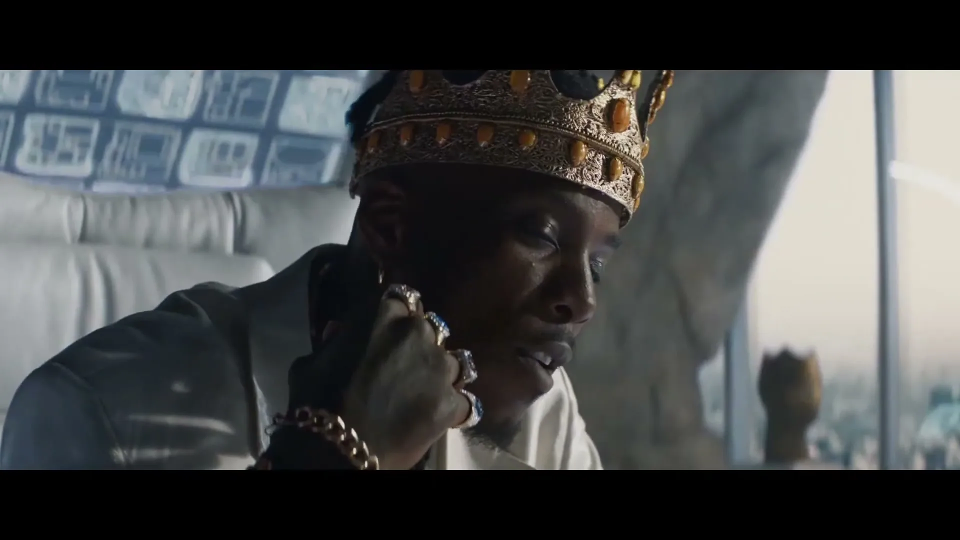 A man wearing a gold crown and rings sits on a white chair, looking down and touching his face in a futuristic setting.