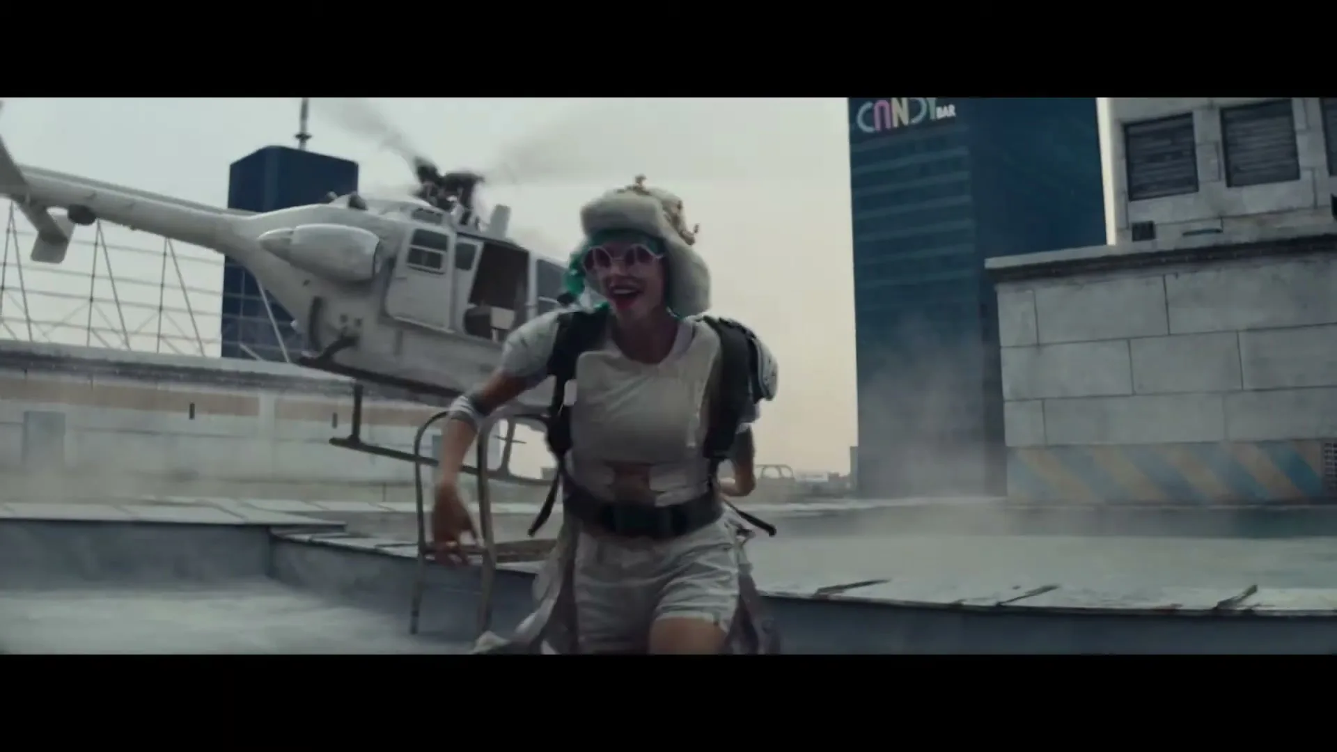 A woman in a white outfit and pink sunglasses runs across a dusty rooftop as a white helicopter takes off behind her, with a city skyline in the background.