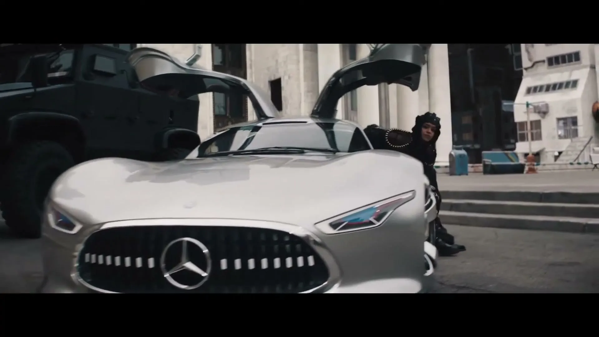 A person with dark skin and dreadlocks, wearing a black outfit, exits a silver Mercedes-Benz sports car with gull-wing doors in a city street.