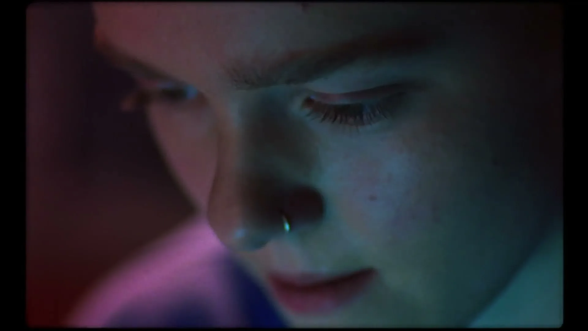 A young person with a nose ring looks down, illuminated by colorful light in a close-up shot.