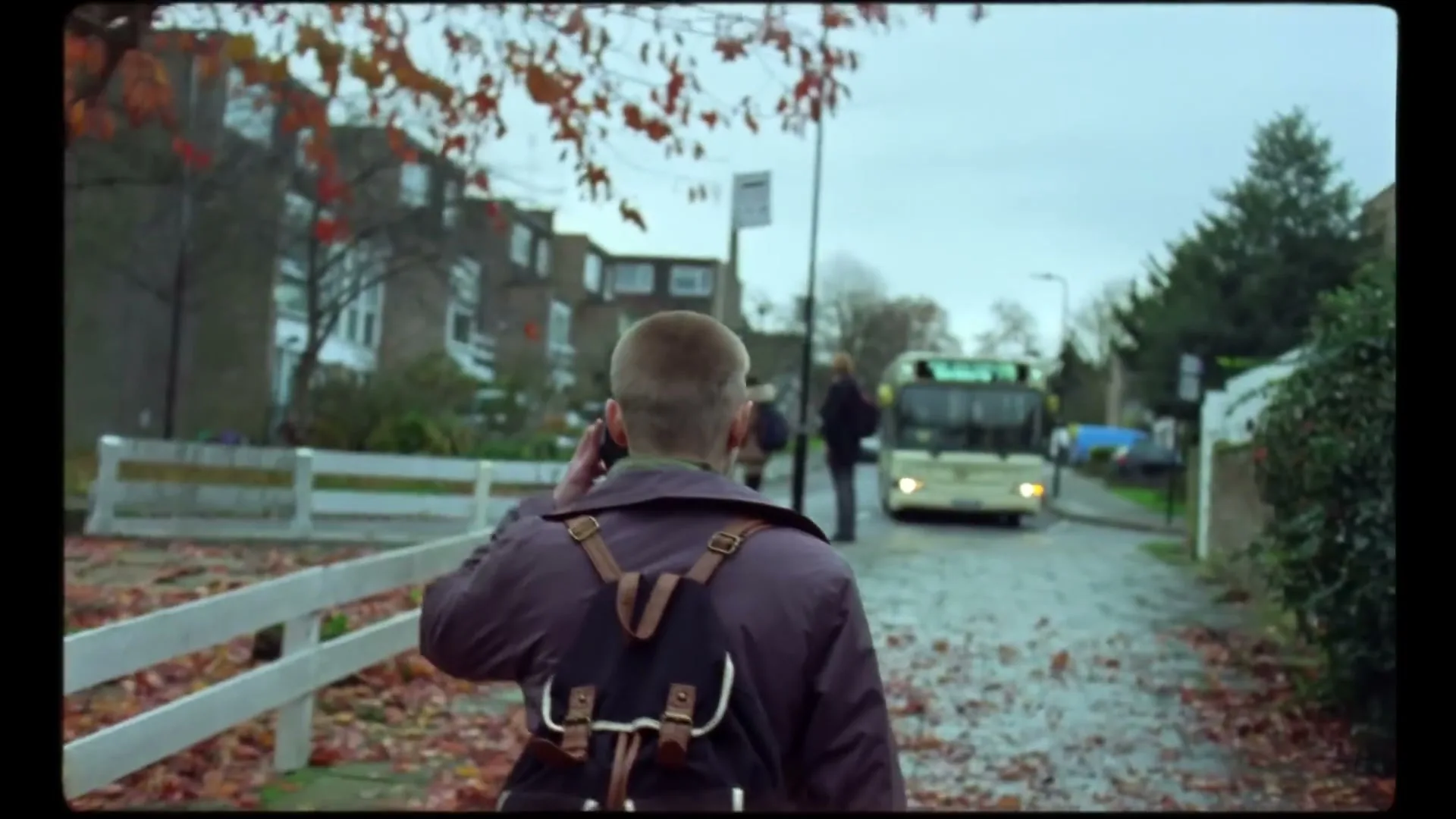 A young man with a backpack walks down a leaf-strewn path, talking on his phone, with a bus and other people in the background on a cloudy day.