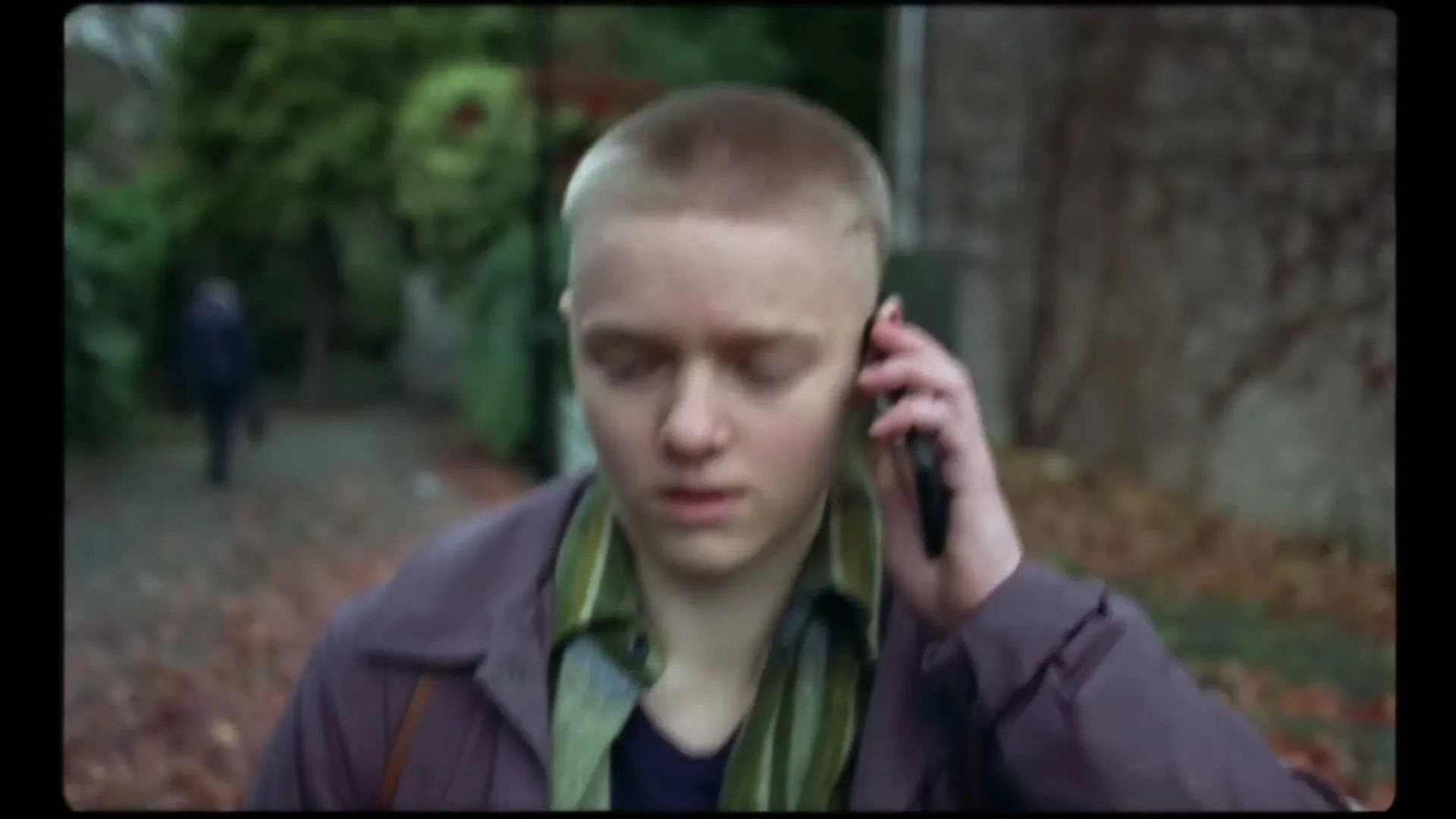 A young person with a buzzcut, wearing a purple jacket and green scarf, walks outdoors while talking on a phone, looking concerned.