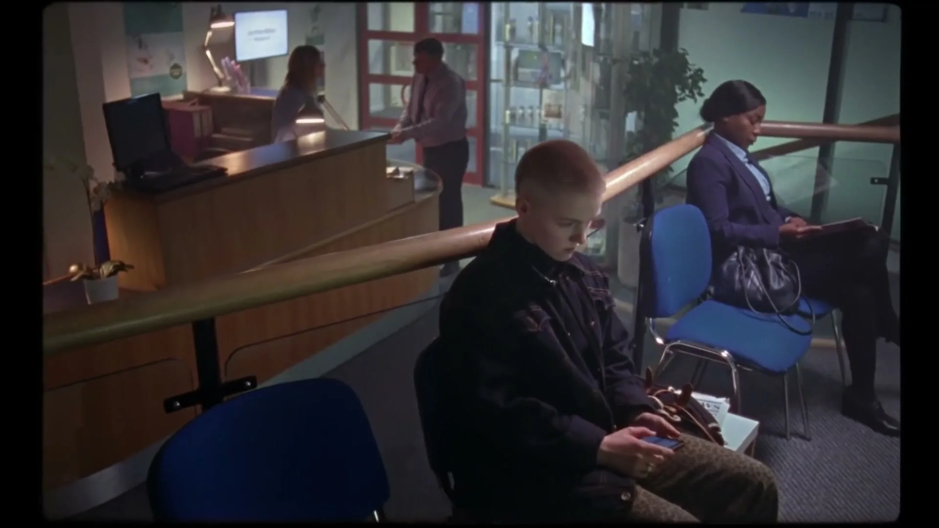 A young person with a buzzcut sits in a waiting room, looking down at their hands, while other people are visible in the background.
