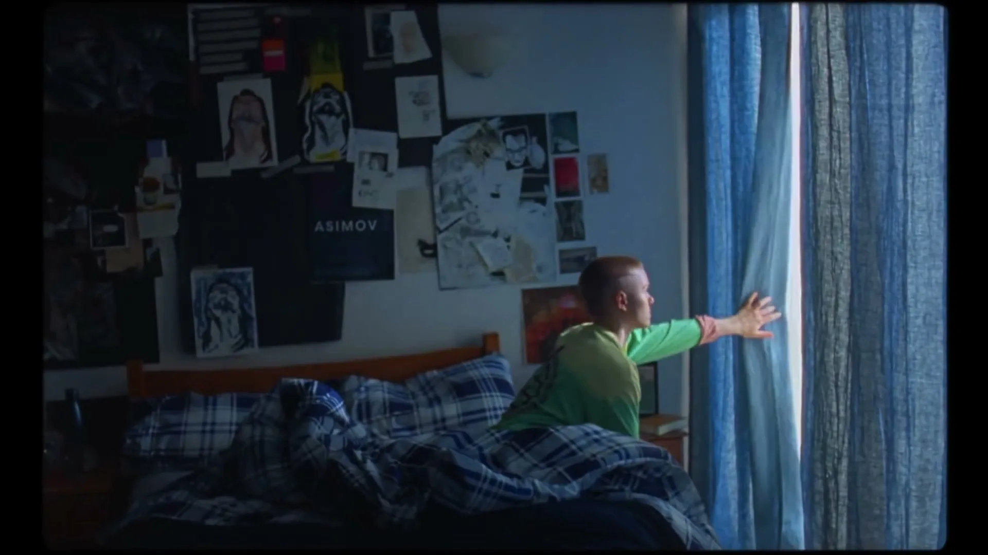 A young person with short hair sits on a bed, reaching out to open blue curtains in a dimly lit bedroom.