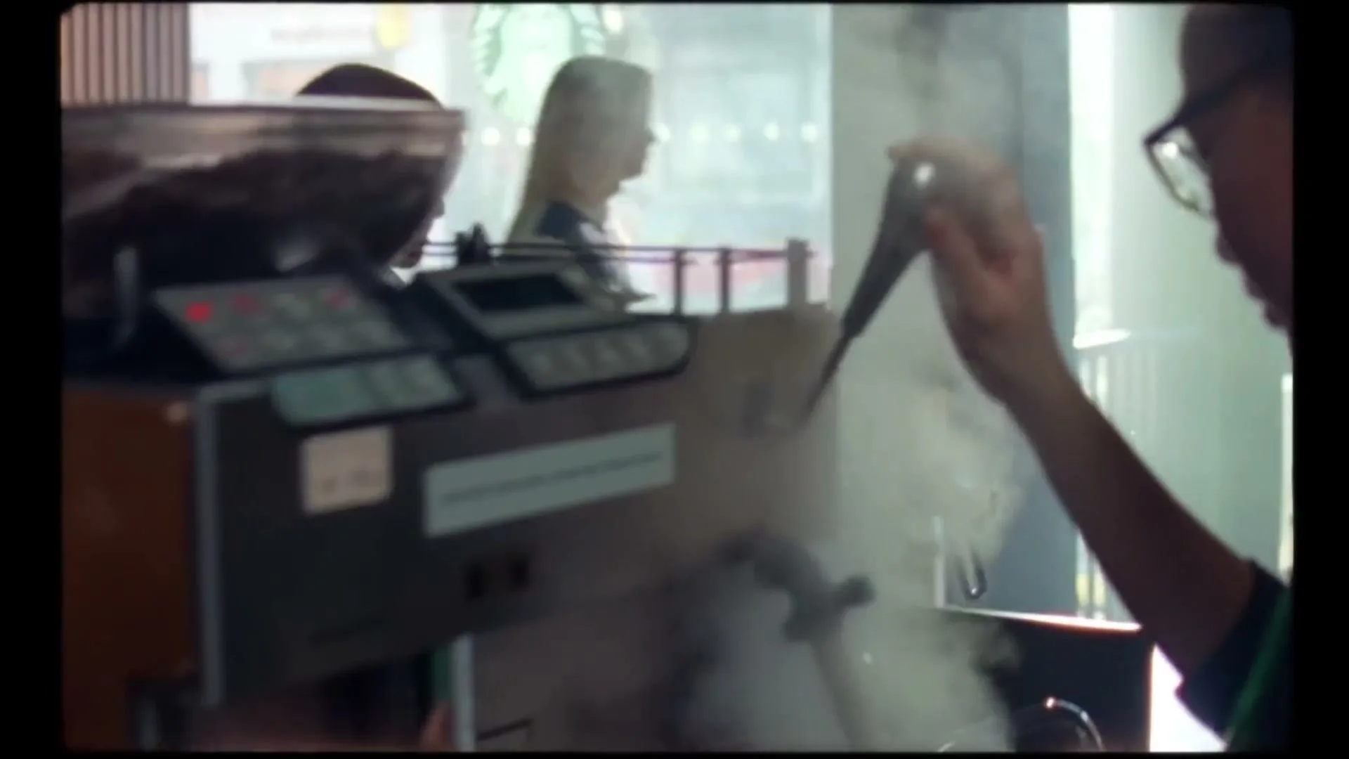 A barista steams milk at an espresso machine, with steam rising, inside a bustling coffee shop during the day.