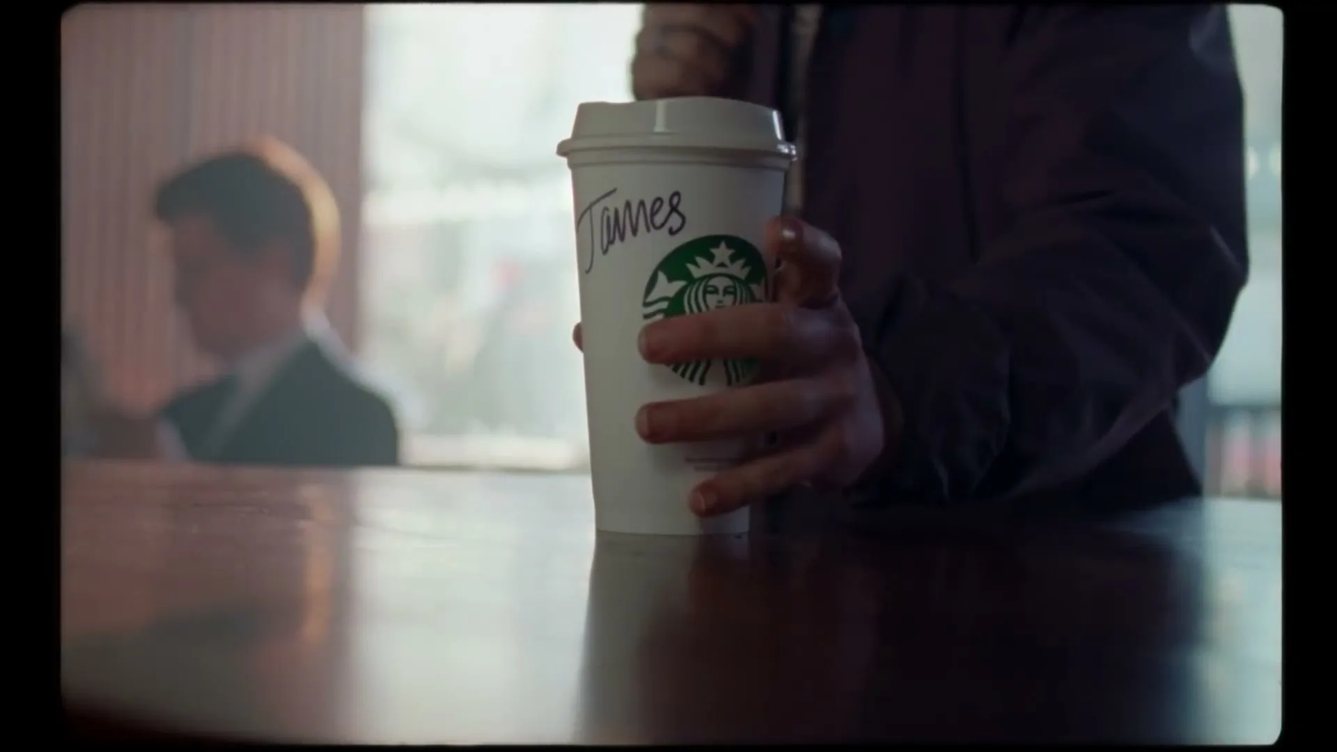 A person's hand reaches for a Starbucks cup with "James" written on it, on a wooden table in a cafe.