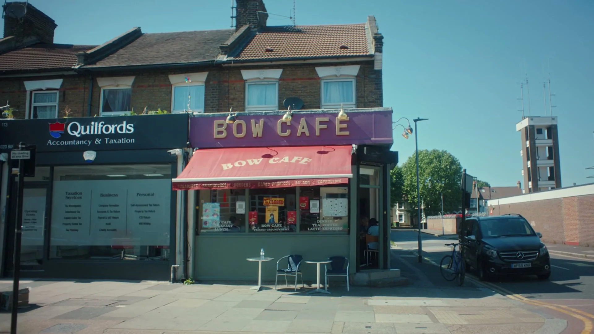 A wide shot of a bustling street in East London, featuring a cafe and an accounting office, with cars and a bus passing by on a sunny day.