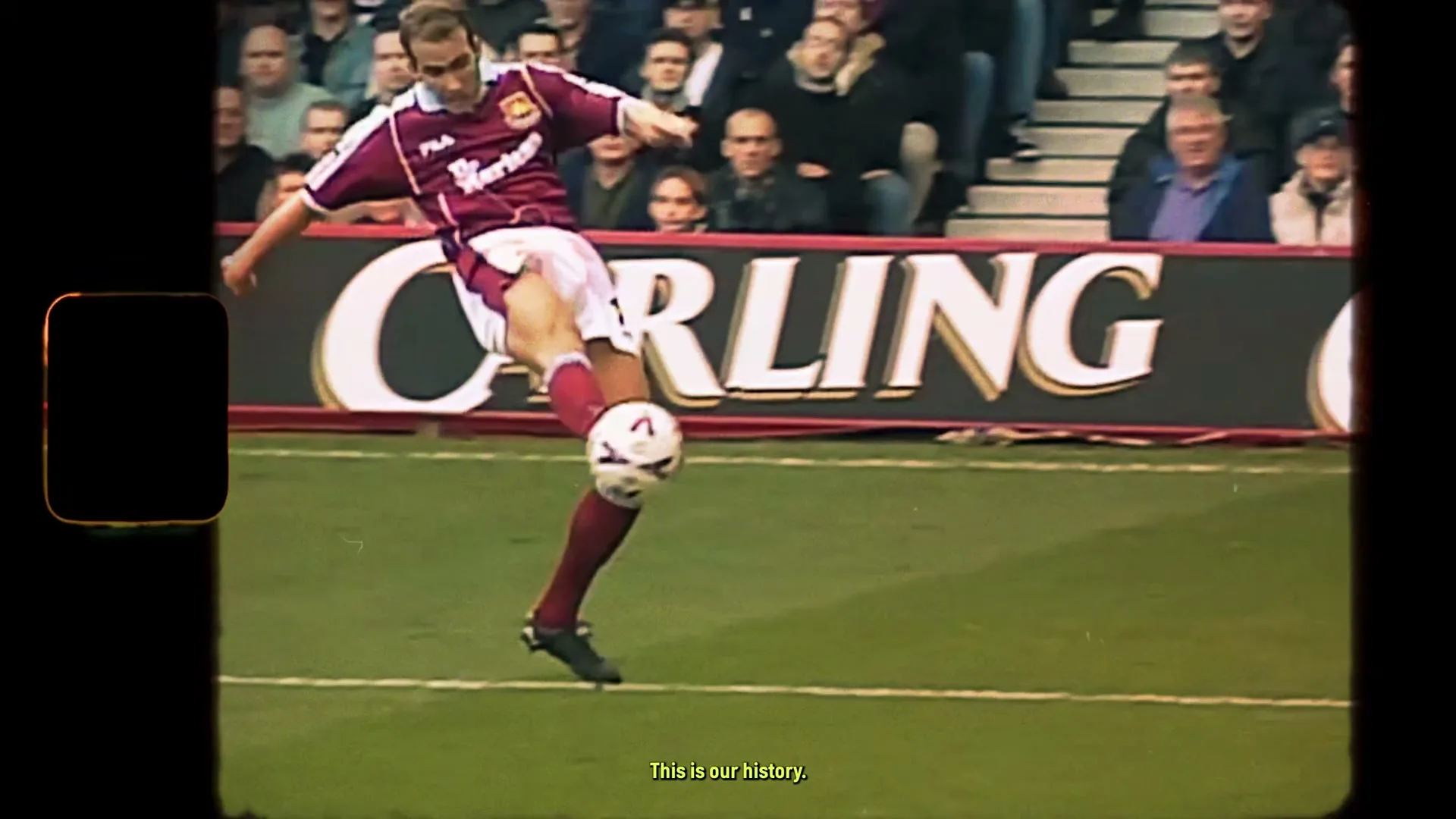 A male soccer player in a maroon and white uniform kicks a soccer ball on a green field in a stadium, celebrating a goal.