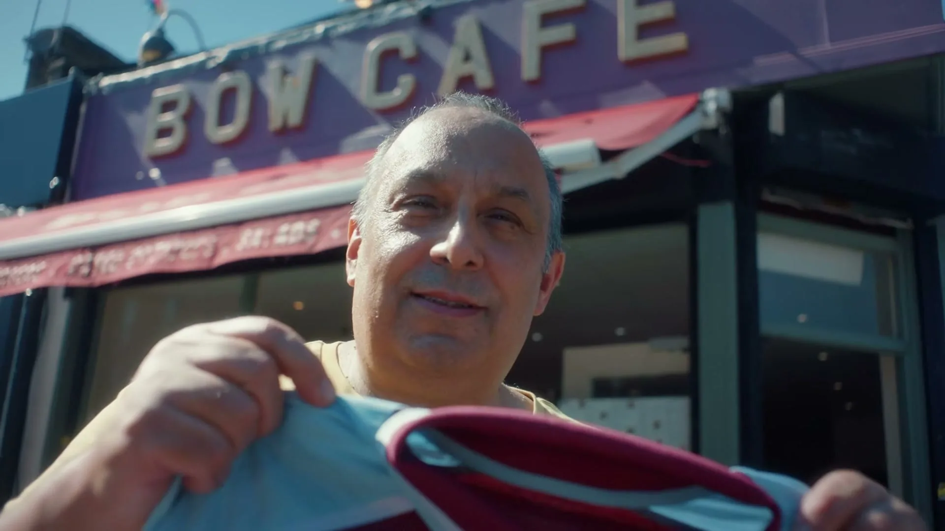 A middle-aged man with a receding hairline smiles at the camera while holding up a West Ham United football jersey in front of the Bow Cafe.