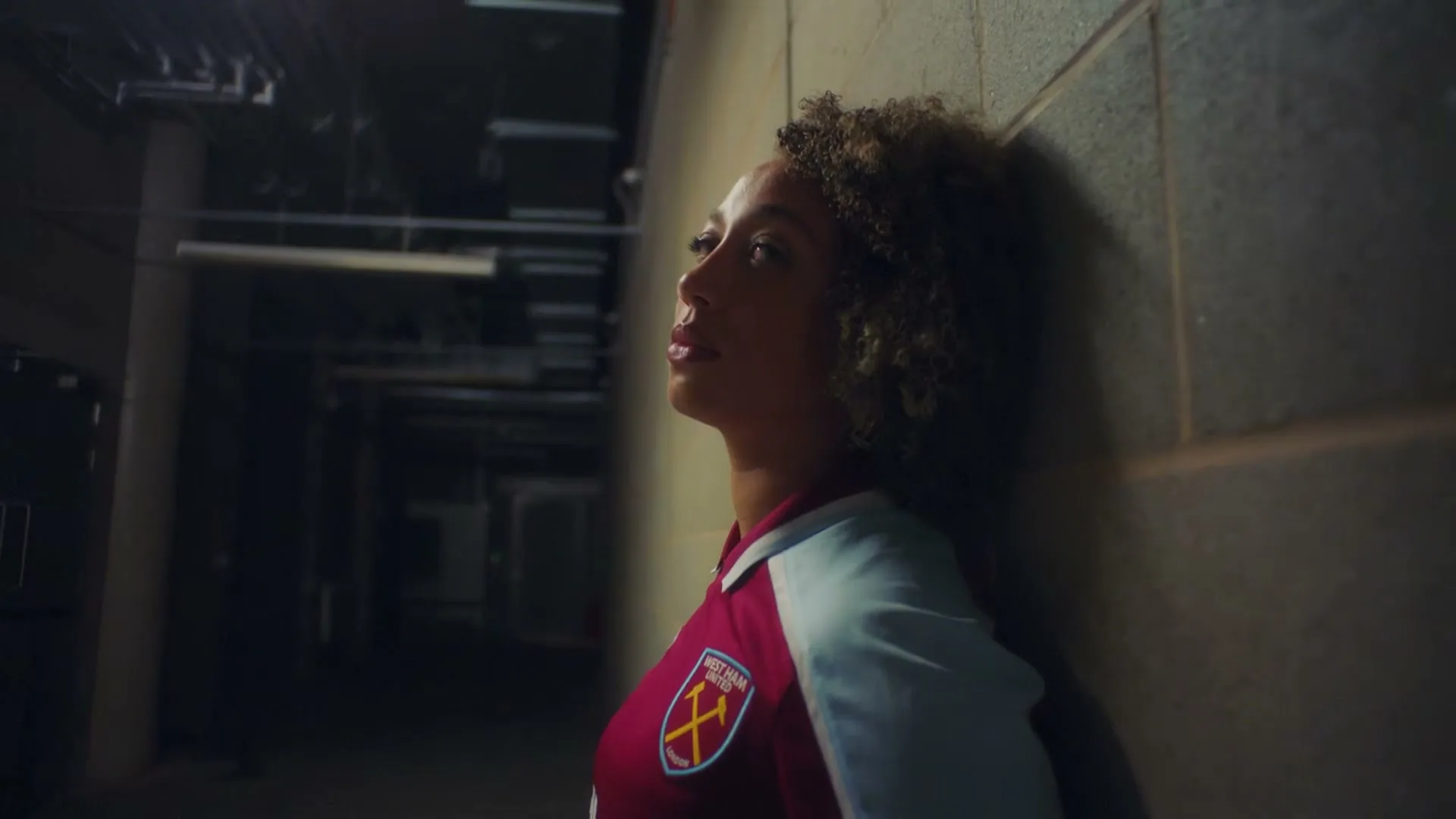 A female soccer player with curly hair, wearing a West Ham United jersey, leans against a concrete wall in a dimly lit stadium tunnel.
