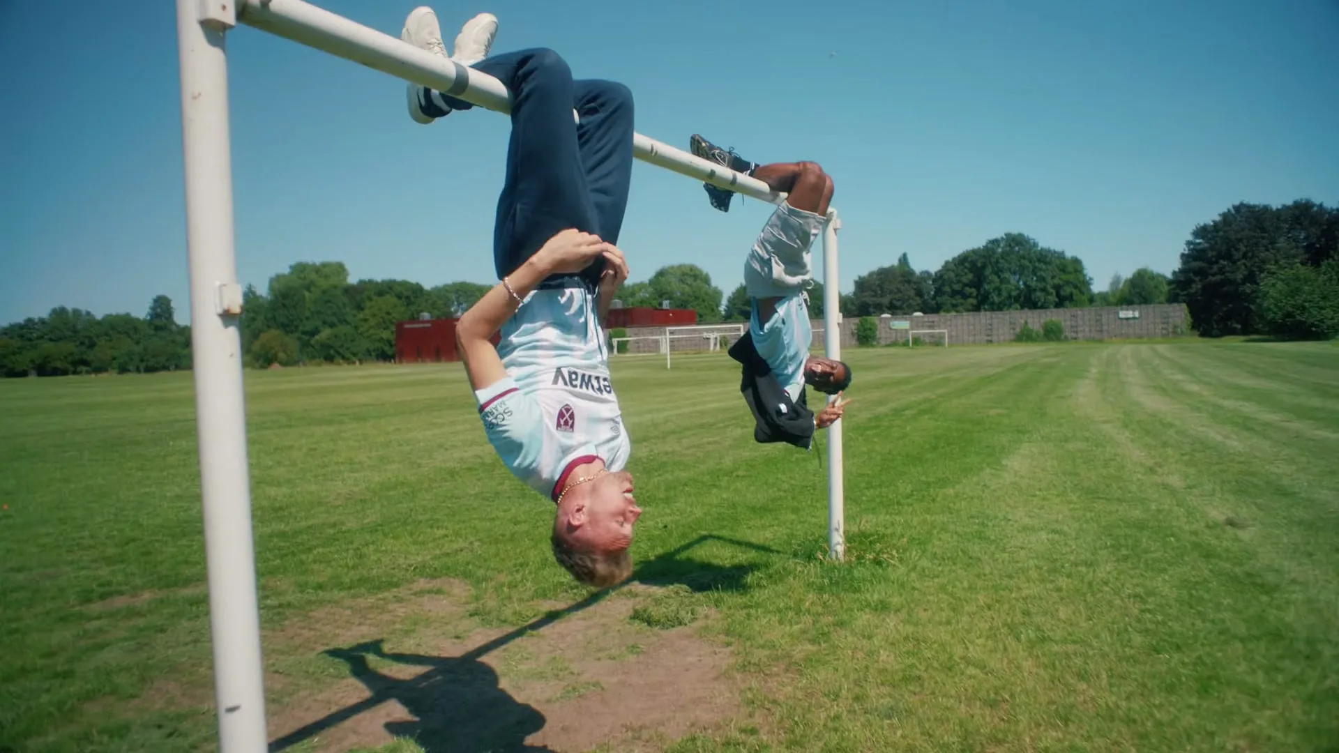 Two young men in West Ham United jerseys hang upside down from a metal bar on a sunny day in a grassy field.