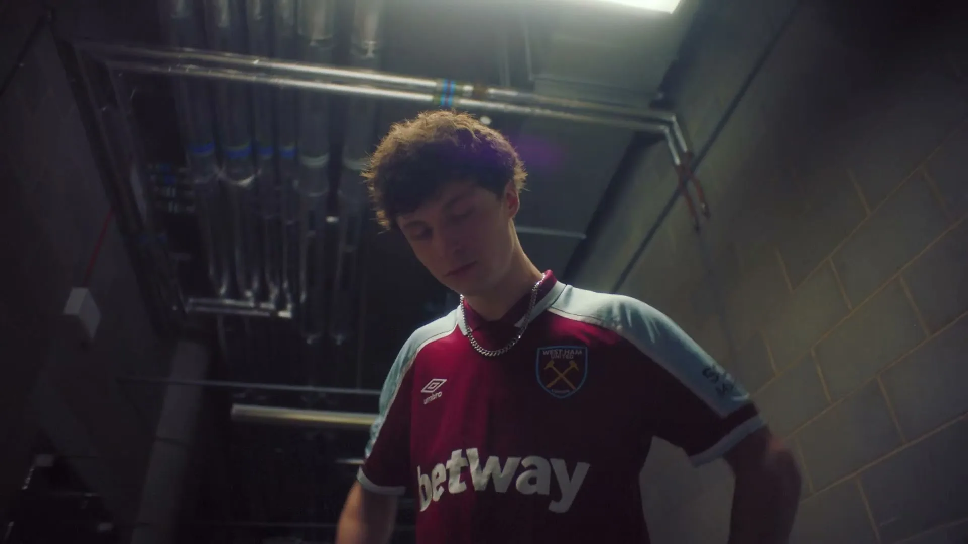 A young male soccer player in a West Ham United jersey looks down then up at the camera in a dark, industrial hallway.