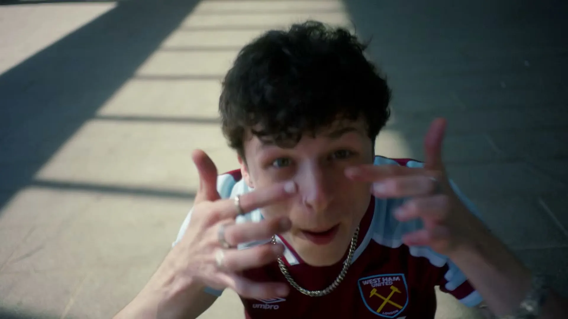 A young man in a West Ham United jersey gestures energetically while looking up at the camera on a sunny day.