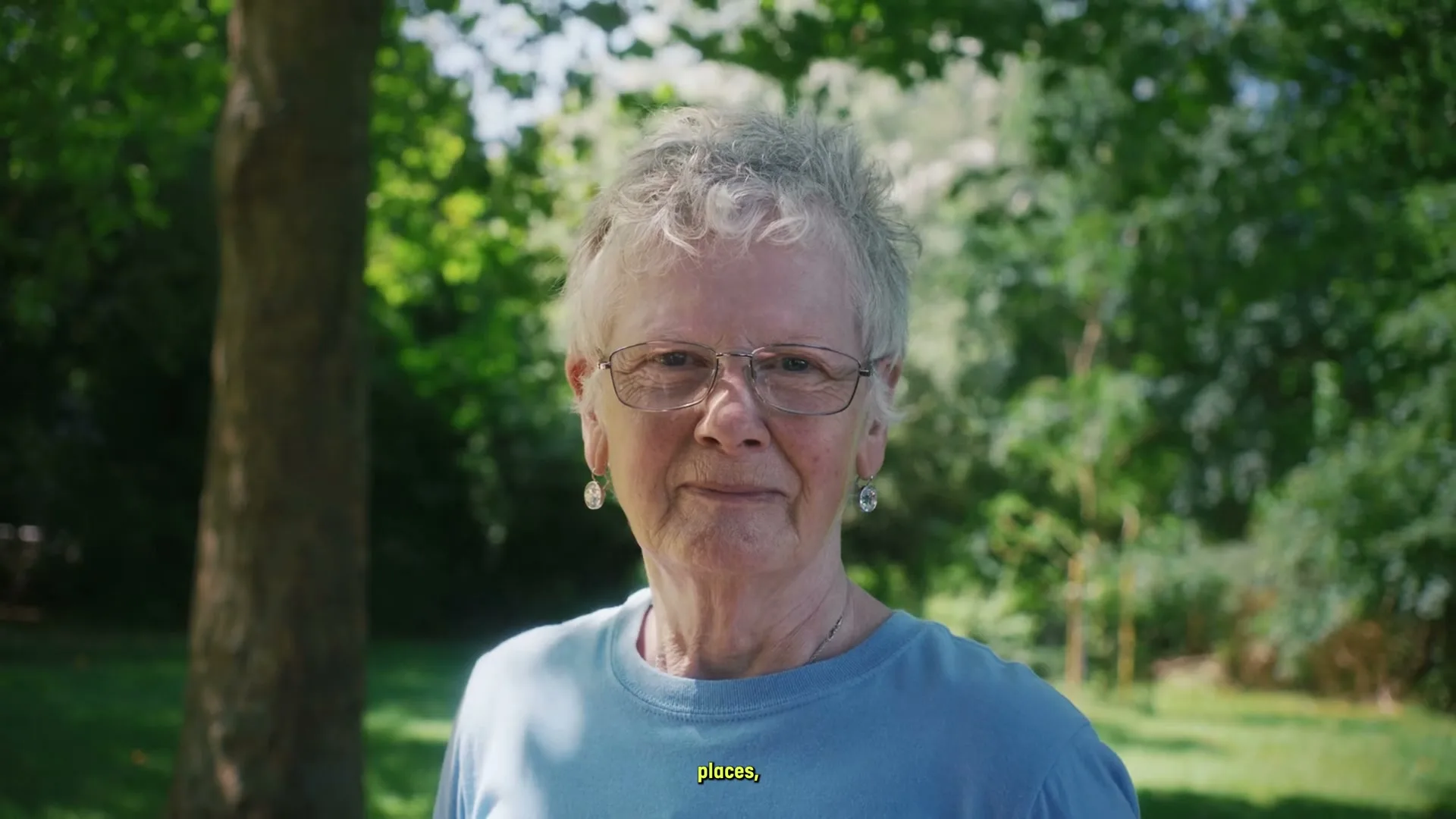 An older woman with short grey hair and glasses smiles gently at the camera, standing outdoors in a park on a sunny day.