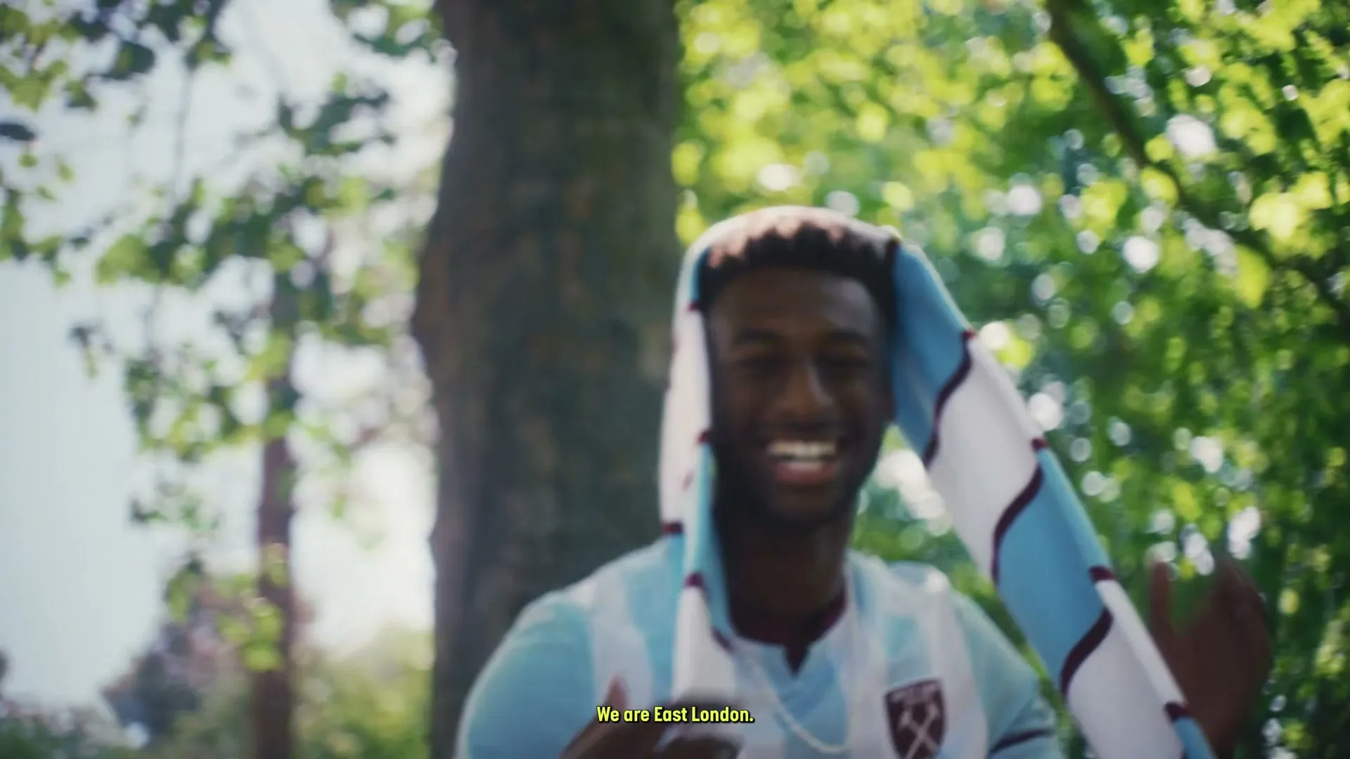 A young Black man in a light blue football jersey and a West Ham United scarf over his head smiles broadly in a park.