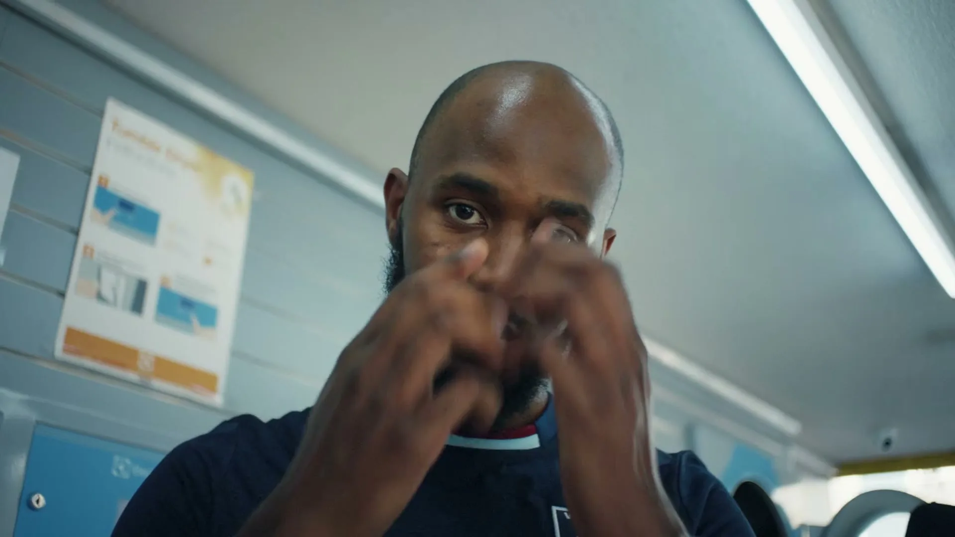 A bald man with a beard, wearing a dark blue t-shirt, looks at the camera and then covers his face with his hands in a laundromat.